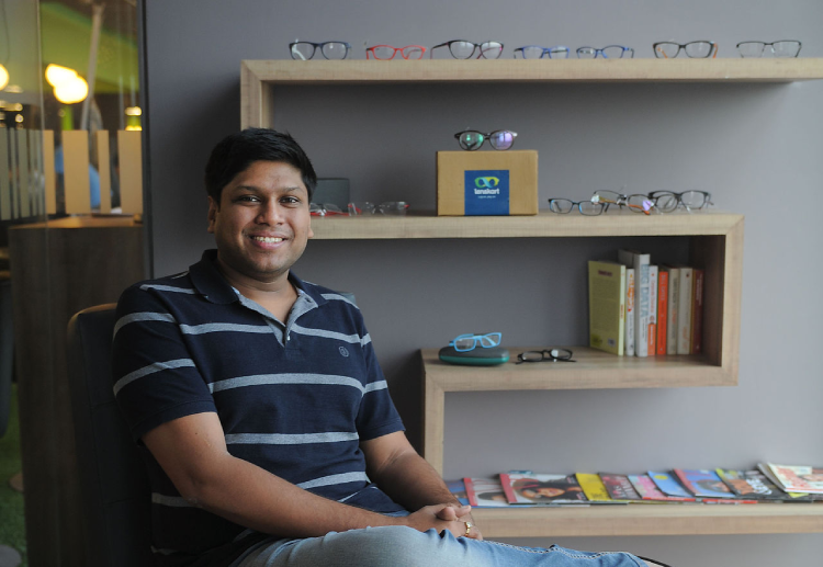 Lenskart founder Peyush Bansal sits by an array of spectacle frames on a shelf 