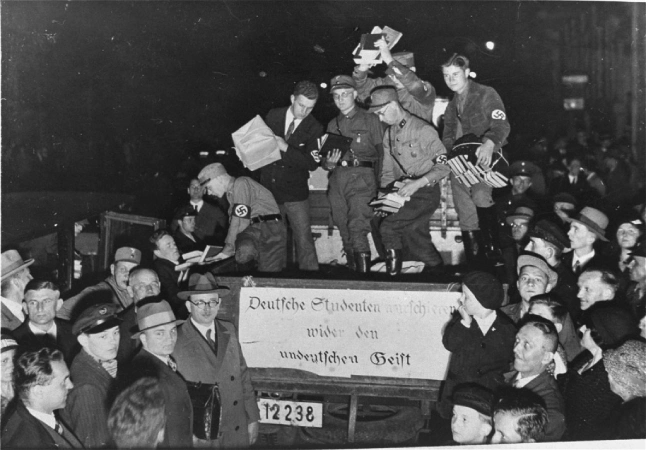 Black-and-white historical photo of Nazi uniformed men throwing books into a pile on a truck while a crowd watches 