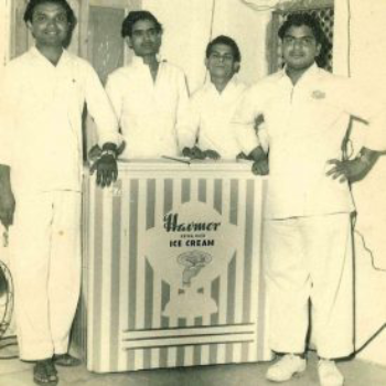 A vintage black-and-white photo showing four men standing around an old ice cream cart, one of them being Satish Chona, with a freezer counter with branding on the front.