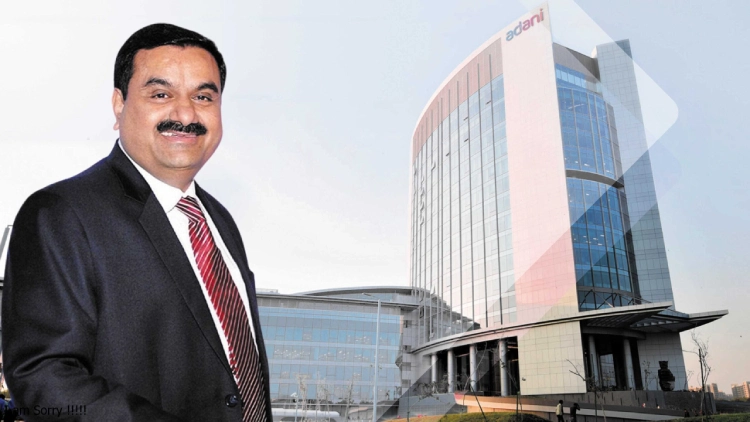 A businessman in a suit standing in front of a modern glass office building with the Adani logo at the top.
